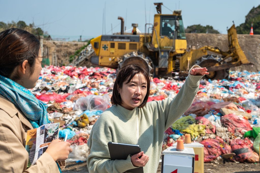 ごみの山を背に埋立処分場の説明をする吉留さん
