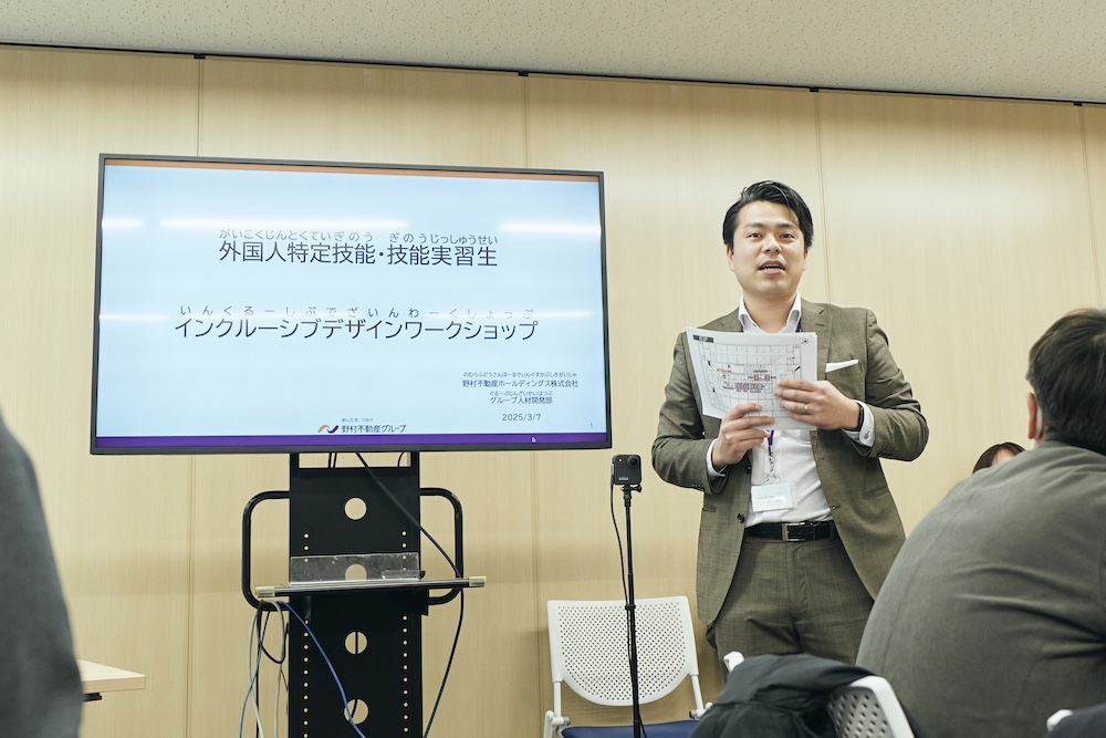 丸山さんが、「外国人特定技能・技能実習生 インクルーシブデザインワークショップ」と映し出されたモニターの横で会場の皆さんに向けて説明している