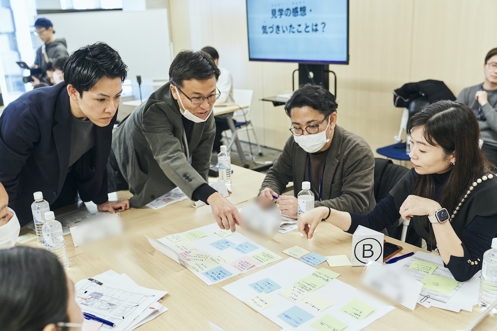 グループワークでテーブルに出された付箋を日本人社員と外国人ワーカーたちが見て、一人の日本人男性社員が指を指して話している