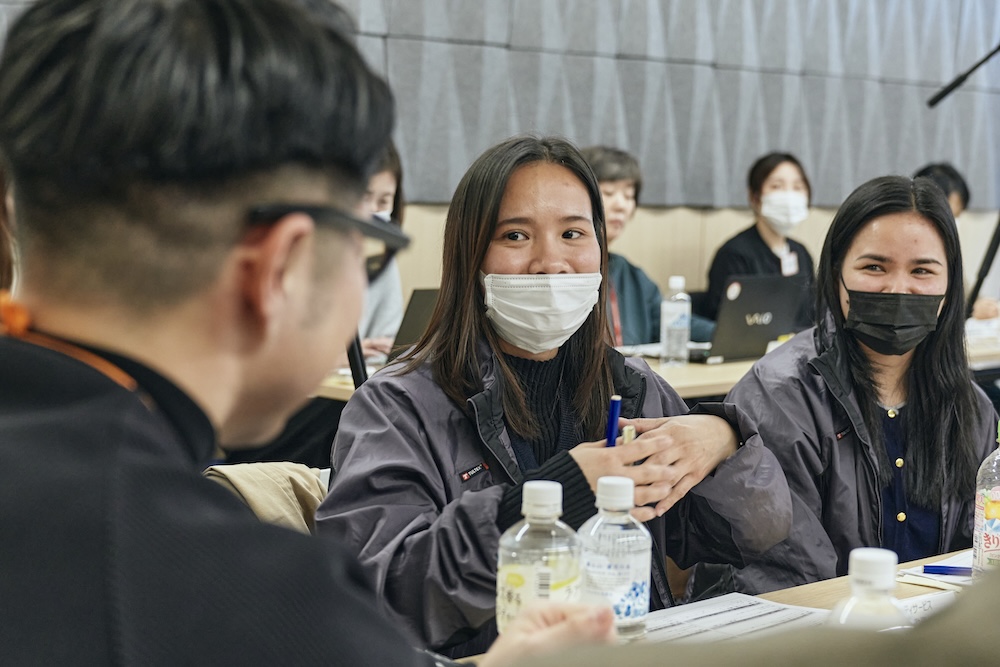 グループワークで、外国人ワーカーの女性が日本人男性社員に向かって話している。もう一人の外国人女性ワーカーが笑顔で見つめる