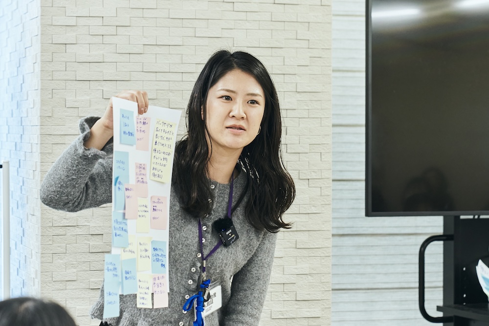 日本人女性社員が付箋の貼られた紙を見せながら全体に向けて説明している