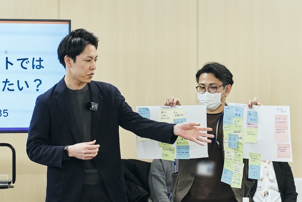 日本人の男性社員2人が付箋の貼られた紙を見せながら全体に向けて説明している