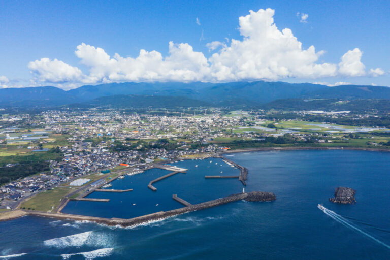 宮崎県都農町の海沿いを上空から望む。防波堤に囲まれた港と町並み、背後には山々が広がる。