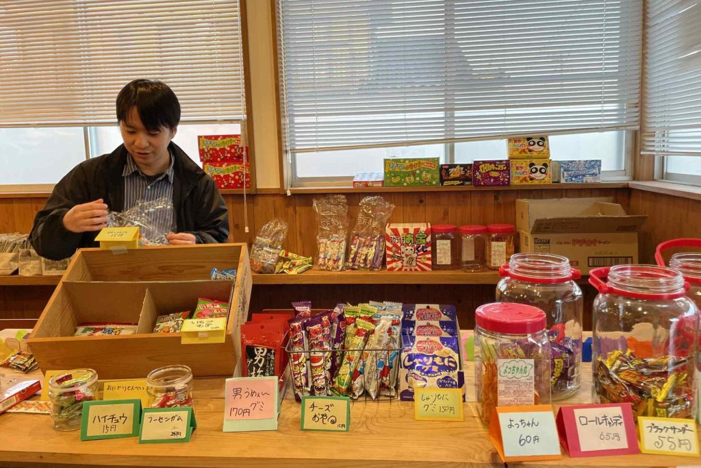 室内の駄菓子屋で、商品の袋詰めをしている様子。カラフルな駄菓子やお菓子の瓶、手書きの値札が並ぶカウンター。