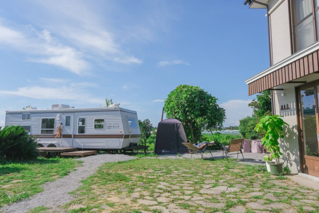 HOSTEL ALAの敷地内に停められたトレーラーとテント。建物の前にはアウトドアチェアが並び、リラックスする人の姿も見える。