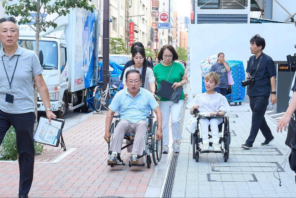 最寄り駅からBFSまでの道を車いすで進み、写真撮影やメモを取りながらルートを確認する参加者たち