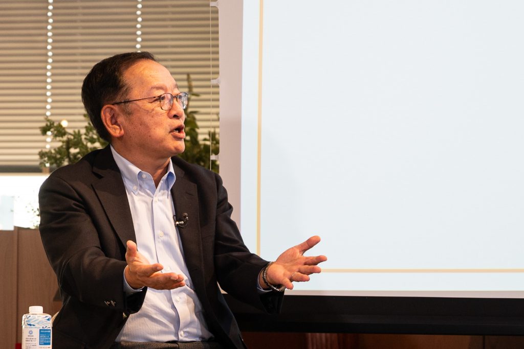 新井グループCEOが両手を広げ、言葉を強調しながら語る様子。横にはスライドが映っている