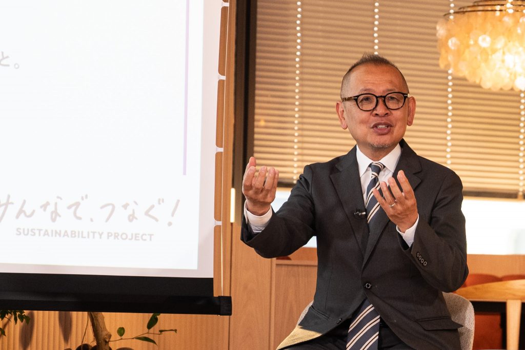 Group CEO Arai speaking with his arms spread wide for emphasis, with a slide displayed next to him
