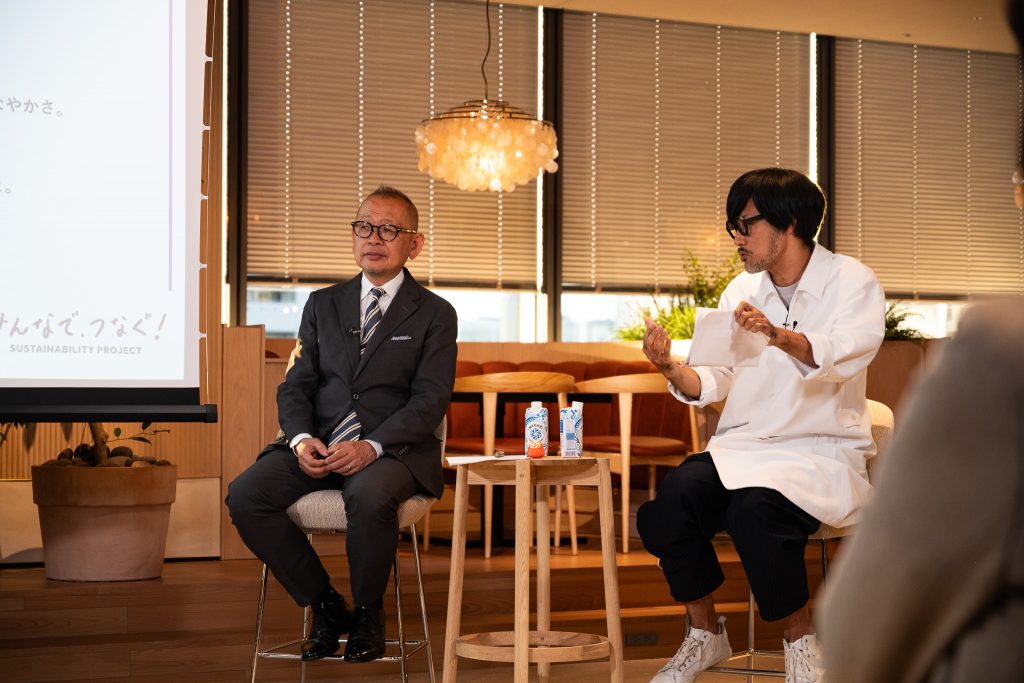 Professor Maeno seated as Mr. Ohata speaks beside him, holding documents.